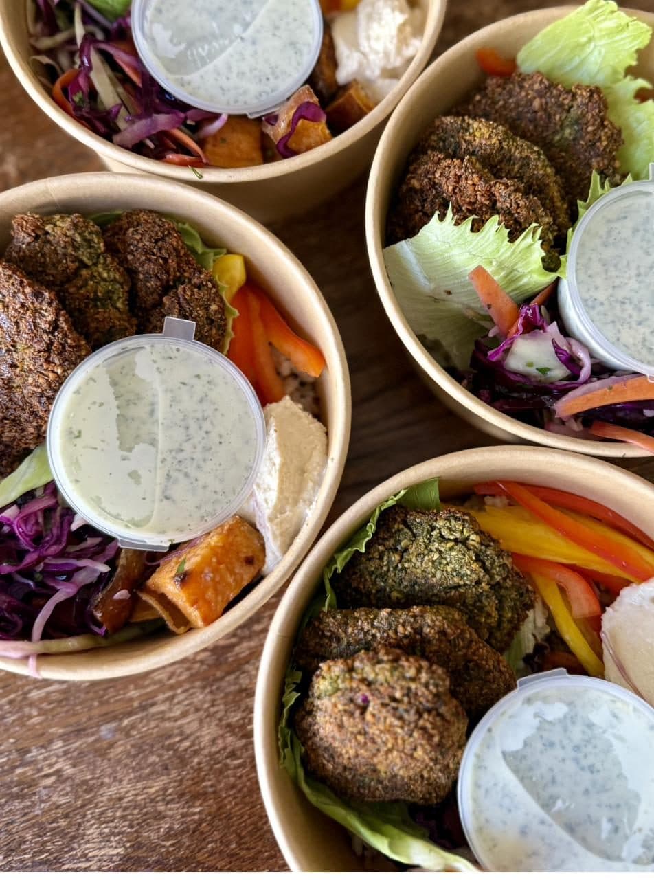 Four Vital Bowl falafel bowls seen from above on a wooden table, each with falafel, hummus, roasted sweet potato, slaw and a pot of green goddess dressing.